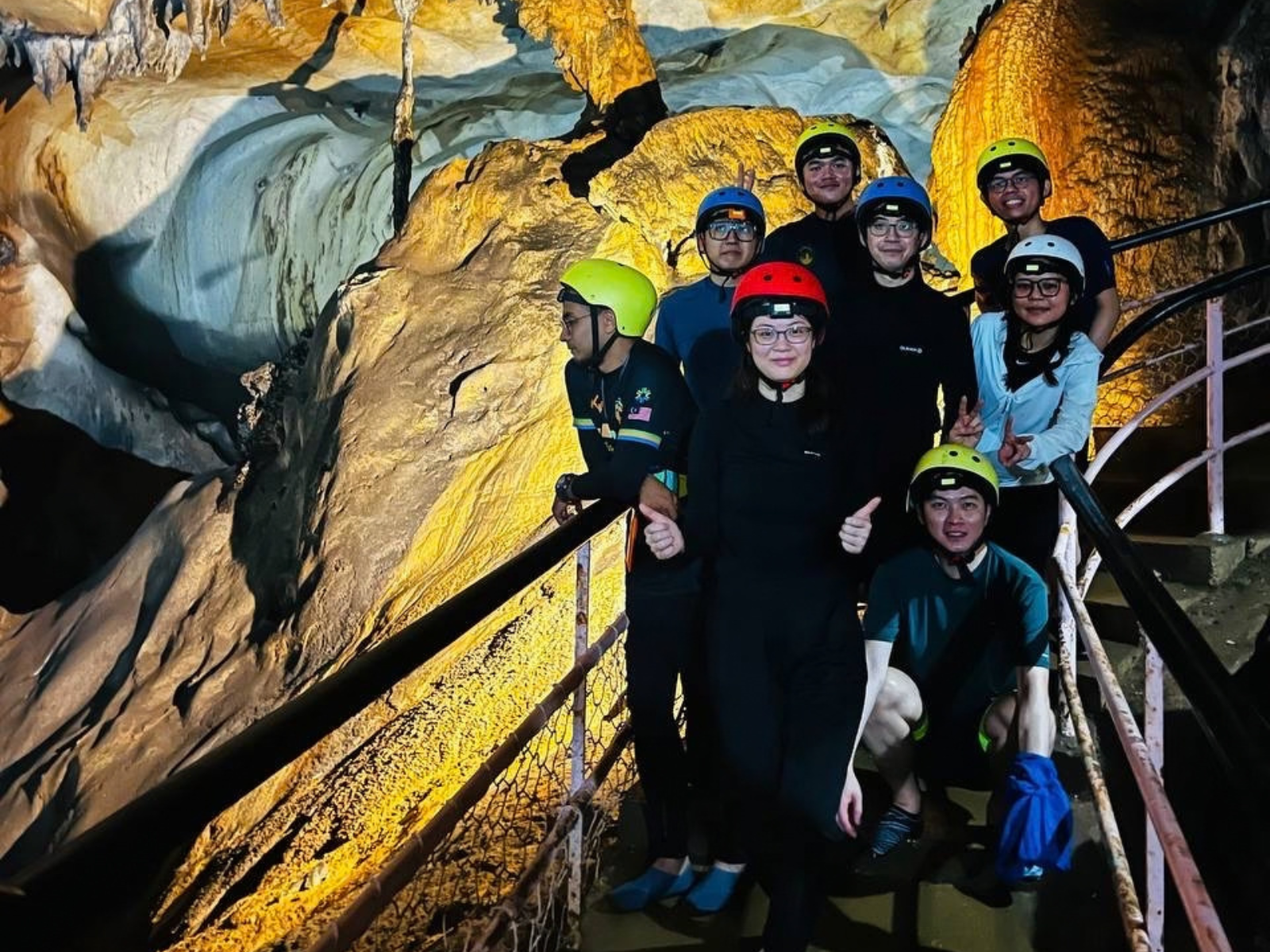 Group in helmets and headlamps posing on a metal staircase inside an illuminated cave passage