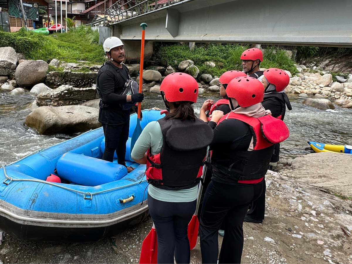 Rafters in red helmets and life jackets listening to a guide beside a blue raft on the riverbank